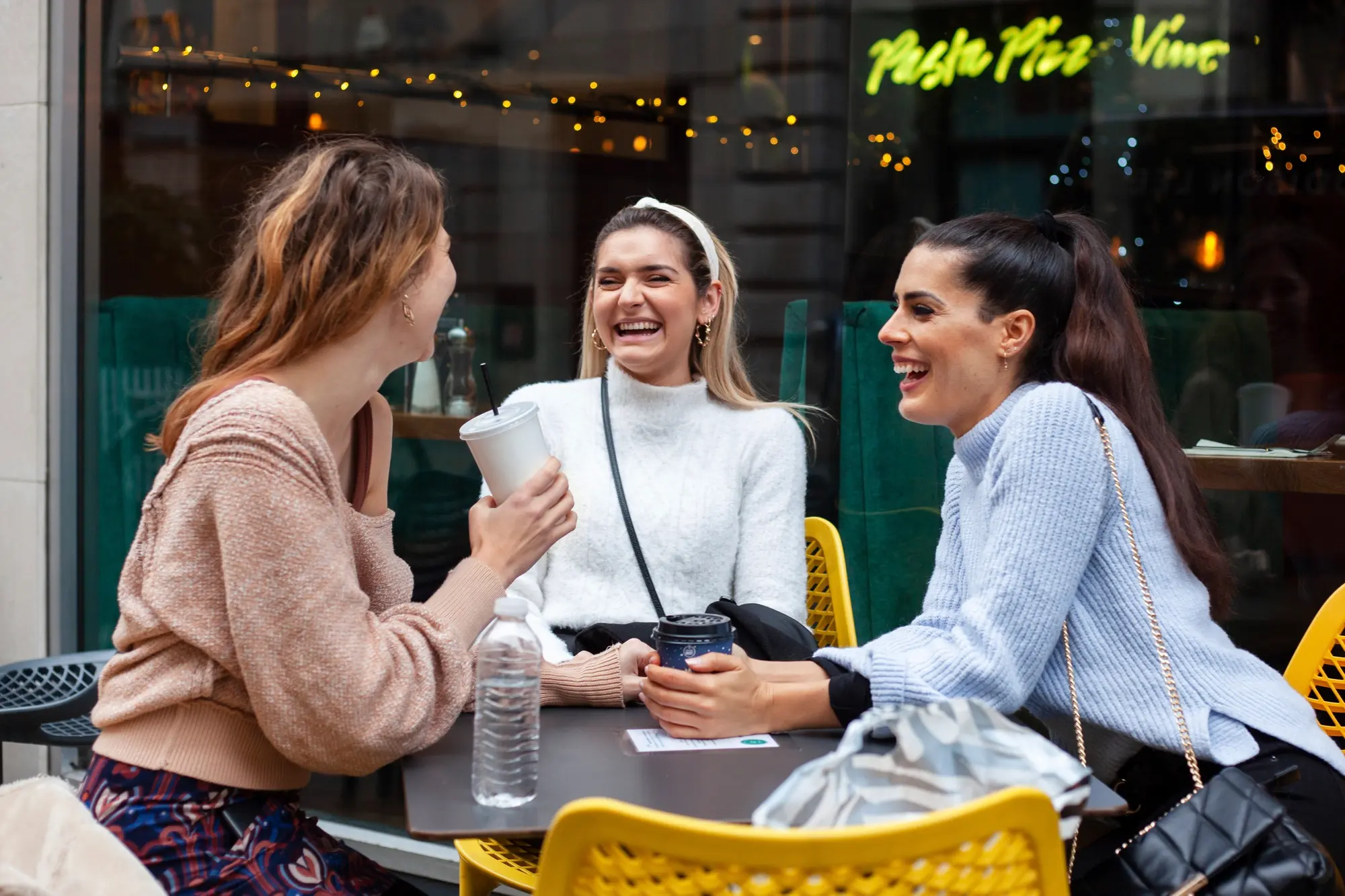 Three women sit at an outdoor table, laughing and holding coffee cups