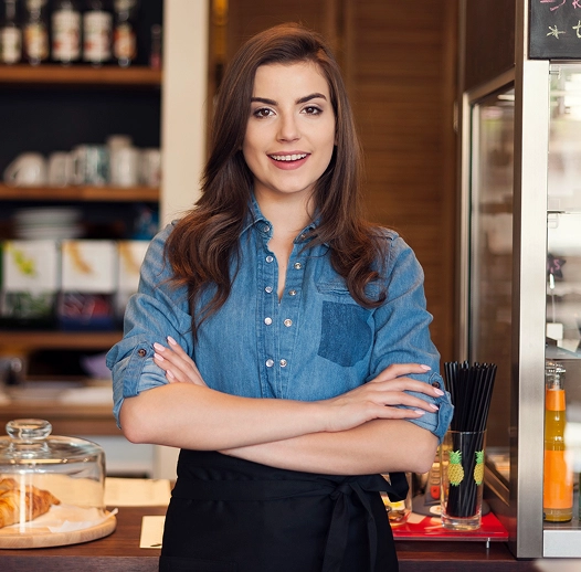 Smiling woman in a denim shirt and black apron stands in a cafe